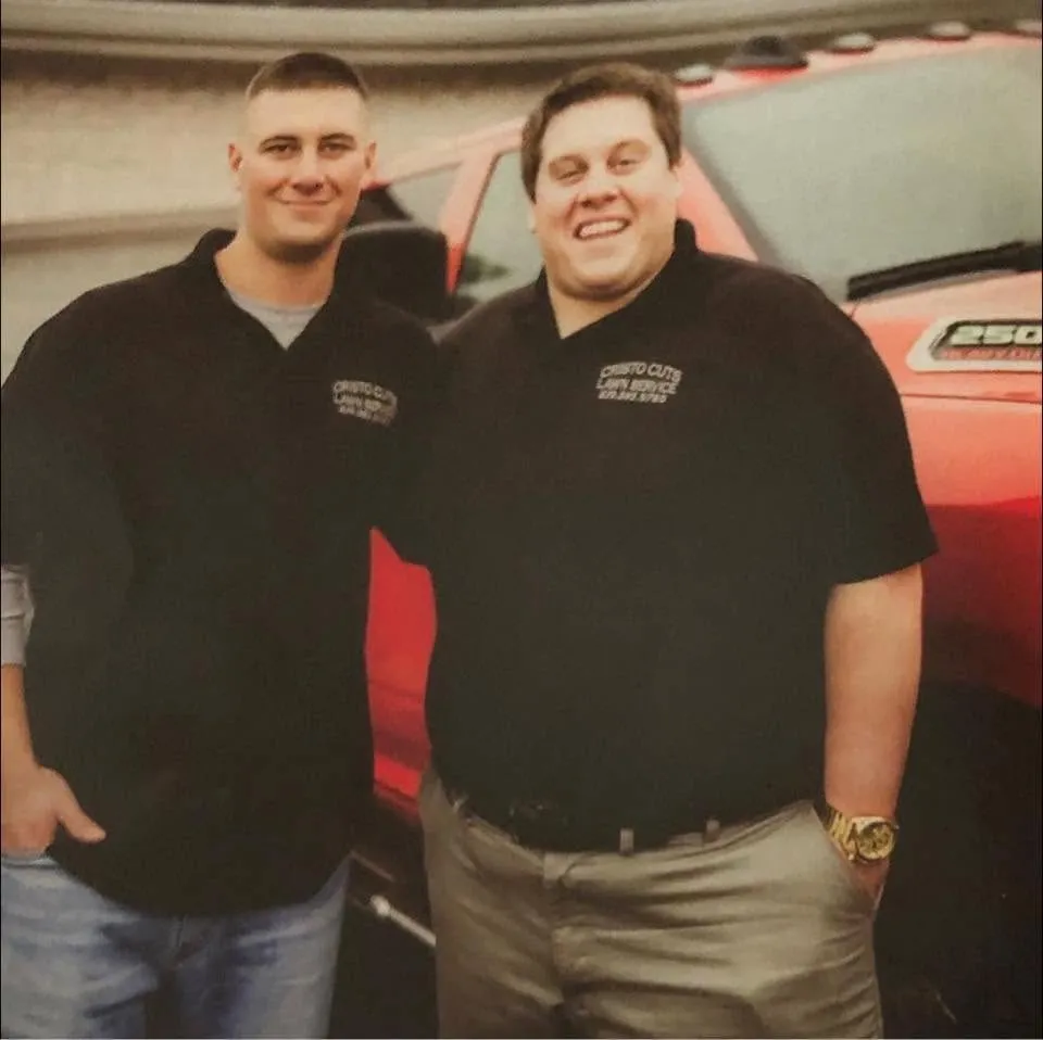 Two members of the Cristo Cuts crew in black company polos in front of a red pickup truck